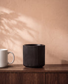Linear Pot in matte black, on a walnut sideboard beside a coffee mug for scale